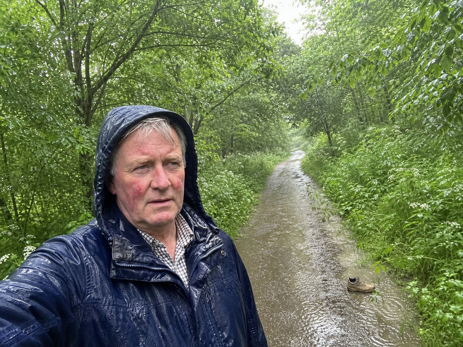 Keith stood on a wet woodland track in a navy waterproof jacket with the hood up, heavy rain falling, a boot visible in the ditch behind him